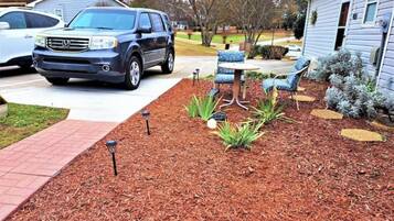 A residential driveway with a parked SUV, a patio area with outdoor furniture, and a landscaped garden bed.
