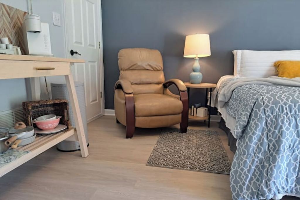 Una camera da letto con un letto, una sedia, un tavolino con una lampada, uno scaffale con oggetti da cucina e un tappeto decorativo.