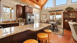 A kitchen with granite countertops, wooden cabinets, a refrigerator, and a dining area with a table and chairs. - Cabin with Long Range Views, Hot Tub, Game Room (Beech Mountain)