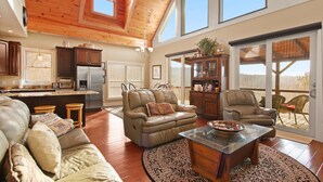 A spacious living area with a high ceiling, wooden beams, a large window, and a sliding glass door leading to an outdoor patio. - Cabin with Long Range Views, Hot Tub, Game Room (Beech Mountain)