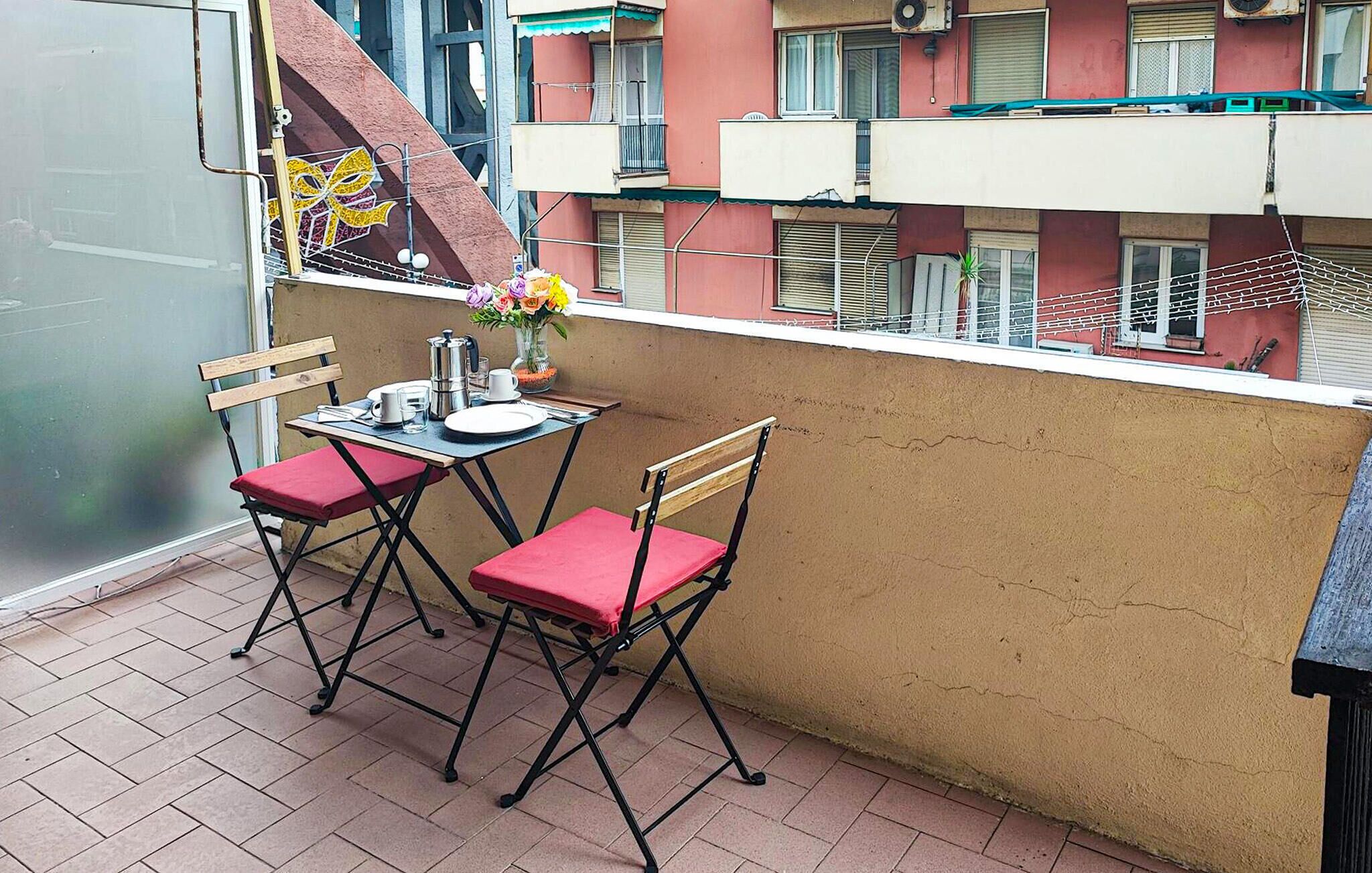 Uma pequena varanda com duas cadeiras dobráveis e uma mesa, um vaso com flores, e vista para um edifício com varandas.