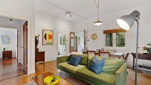 A spacious living room with a green sofa, wooden flooring, a dining area with a table and chairs, and a large window providing natural light. - Blackheath Breeze Cottage - Nature Escape (Blackheath)