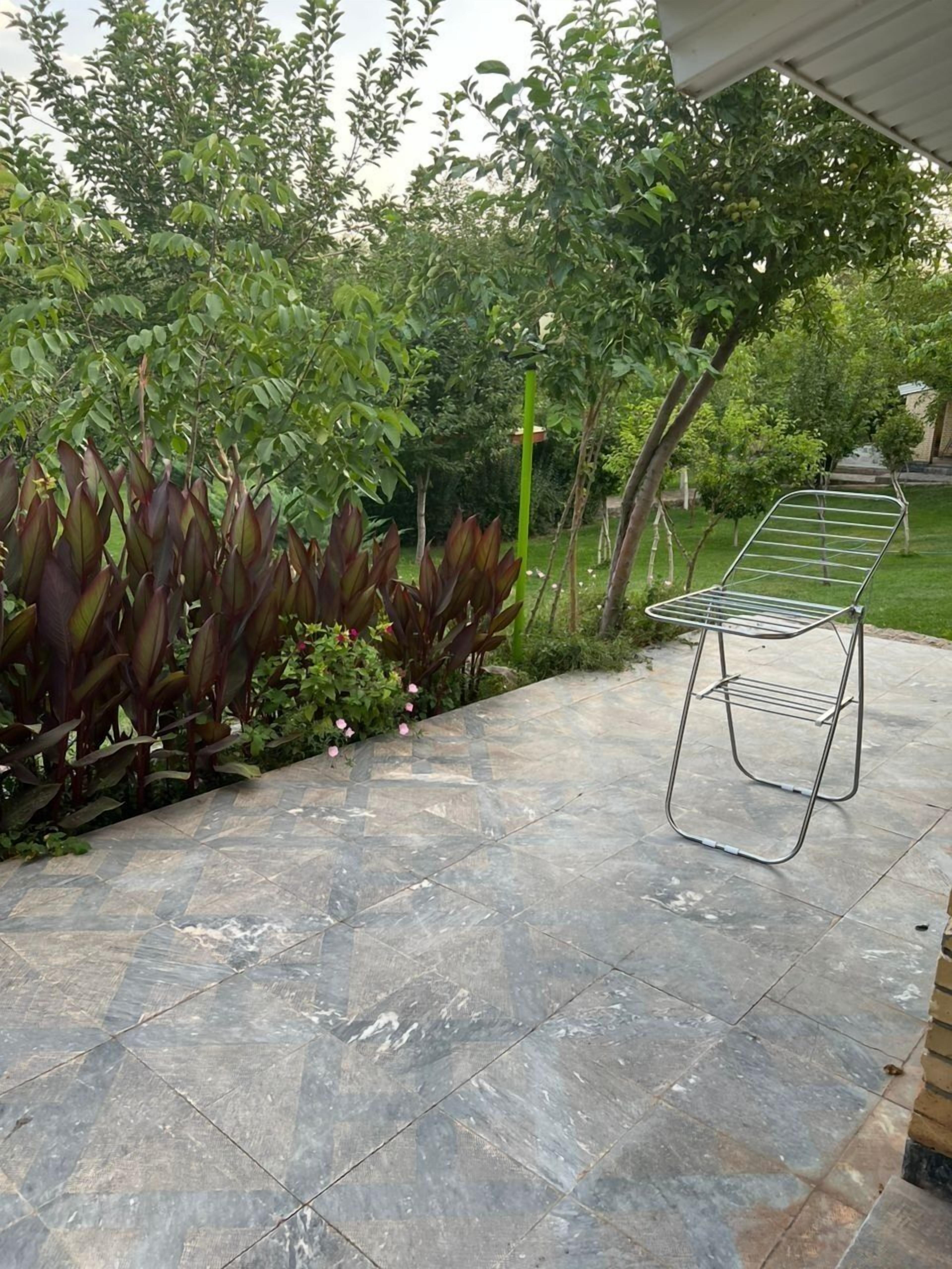 Un patio con una silla de metal, baldosas de piedra y abundante vegetación.
