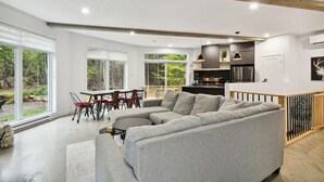 A modern living room with a grey sectional sofa, a dining area with a table and chairs, and a kitchen with a black countertop and stainless steel appliances.