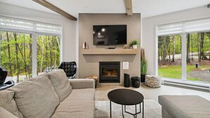 A modern living room with a fireplace, a flat-screen TV mounted above it, and large windows offering a view of trees.