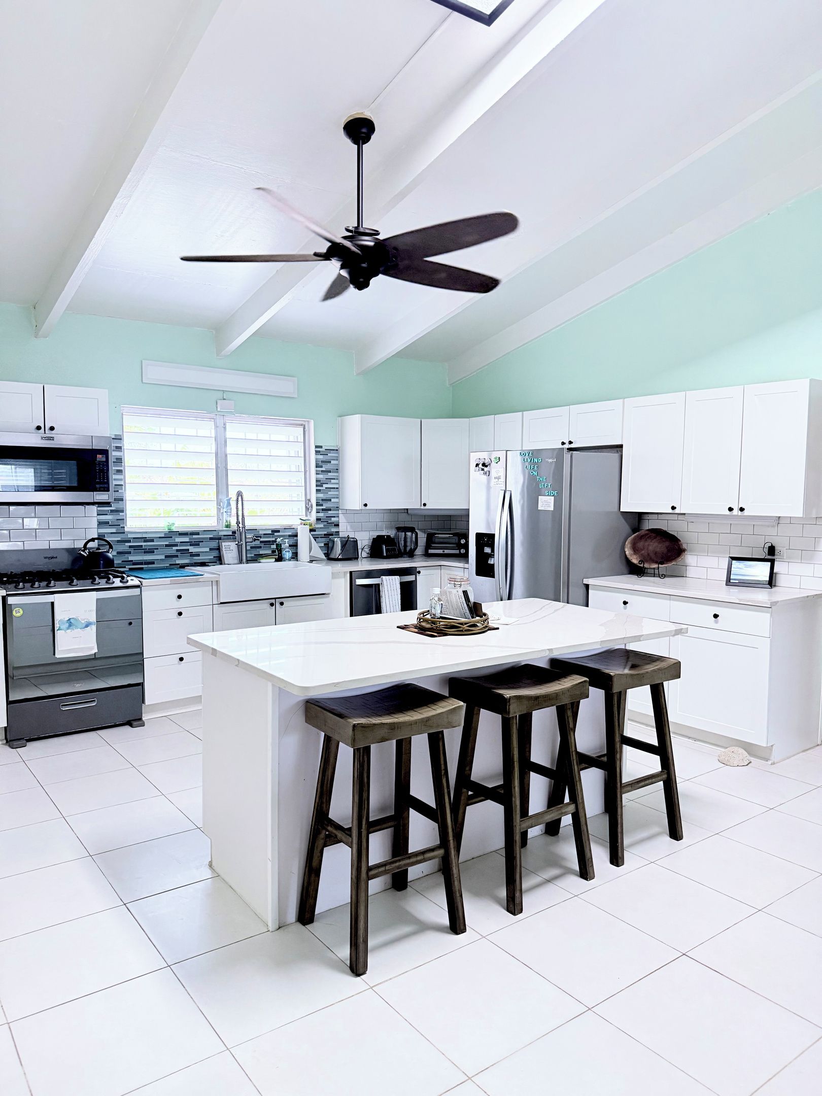 Une cuisine moderne avec des meubles blancs, un îlot central doté de tabourets, un ventilateur de plafond et un réfrigérateur en acier inoxydable.