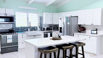 Une cuisine moderne avec des meubles blancs, un îlot central doté de tabourets, un ventilateur de plafond et un réfrigérateur en acier inoxydable.