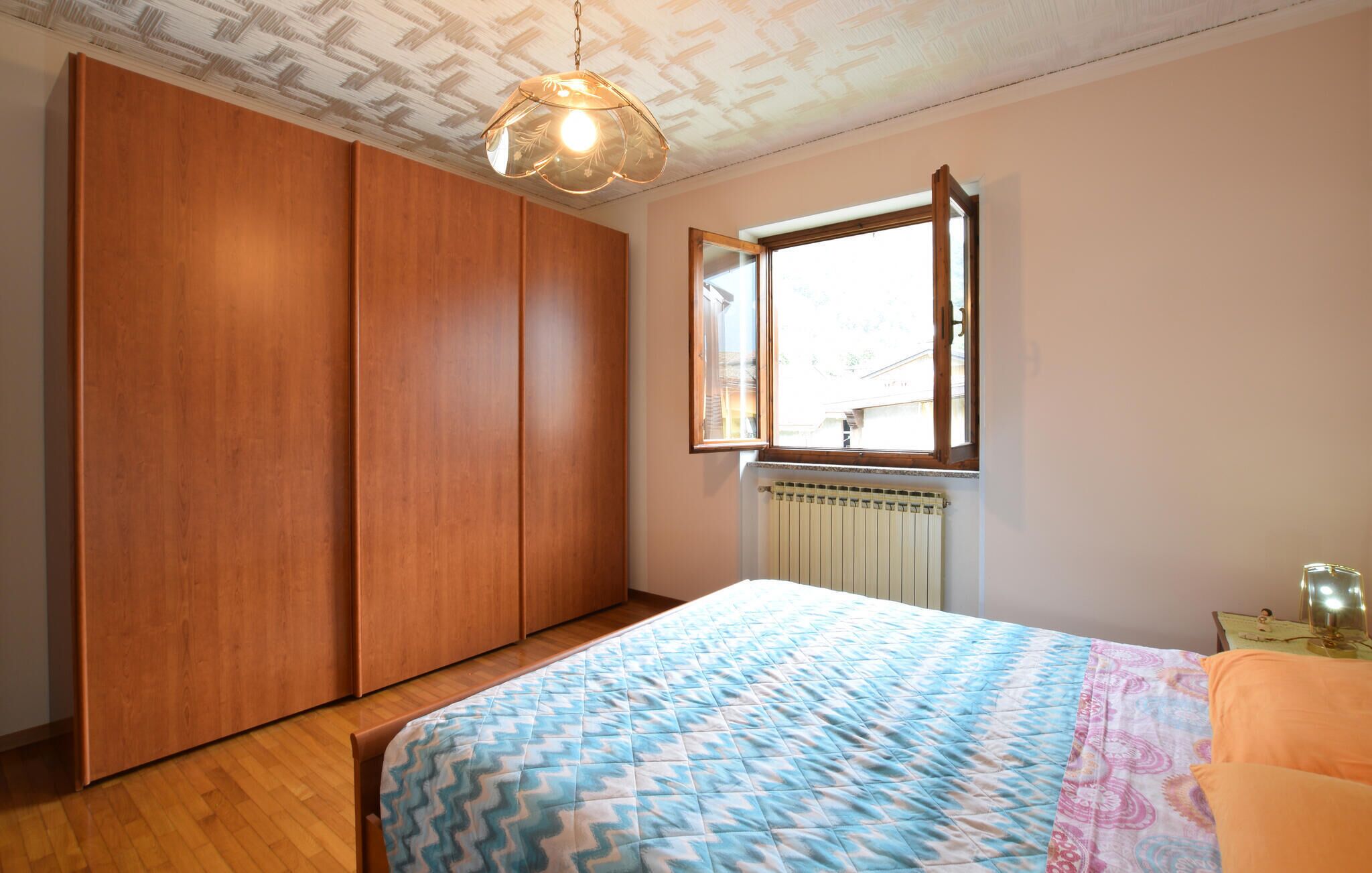 Quarto com armário de madeira, cama com colcha estampada, janela com vista e radiador.