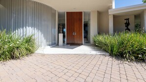 A modern building with a curved facade, a wooden door, and a paved entrance area with tall grasses. - Luxe Hollywood Hills Modern, City Views, 84°F Pool (Los Angeles)