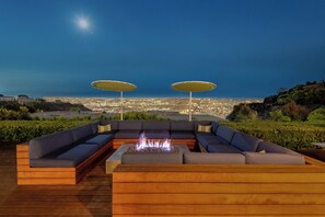 A deck with a fire pit, two umbrellas, and a sofa facing a cityscape at night. - Luxe Hollywood Hills Modern, City Views, 84°F Pool (Los Angeles)