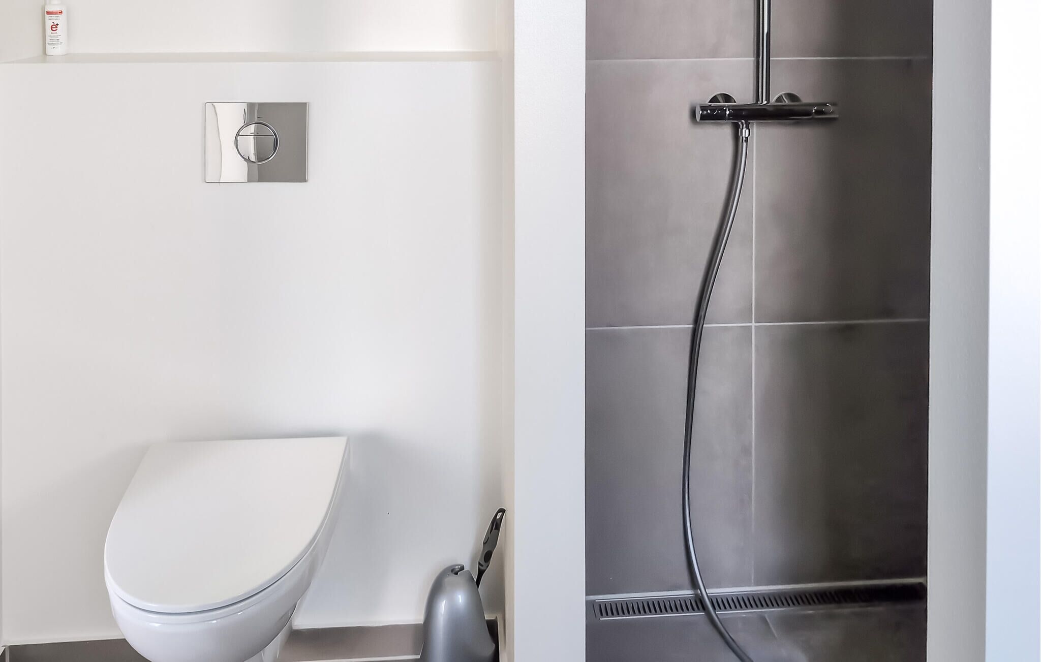 A modern bathroom with a white toilet, a wall-mounted flush handle, and a shower area with a handheld showerhead.