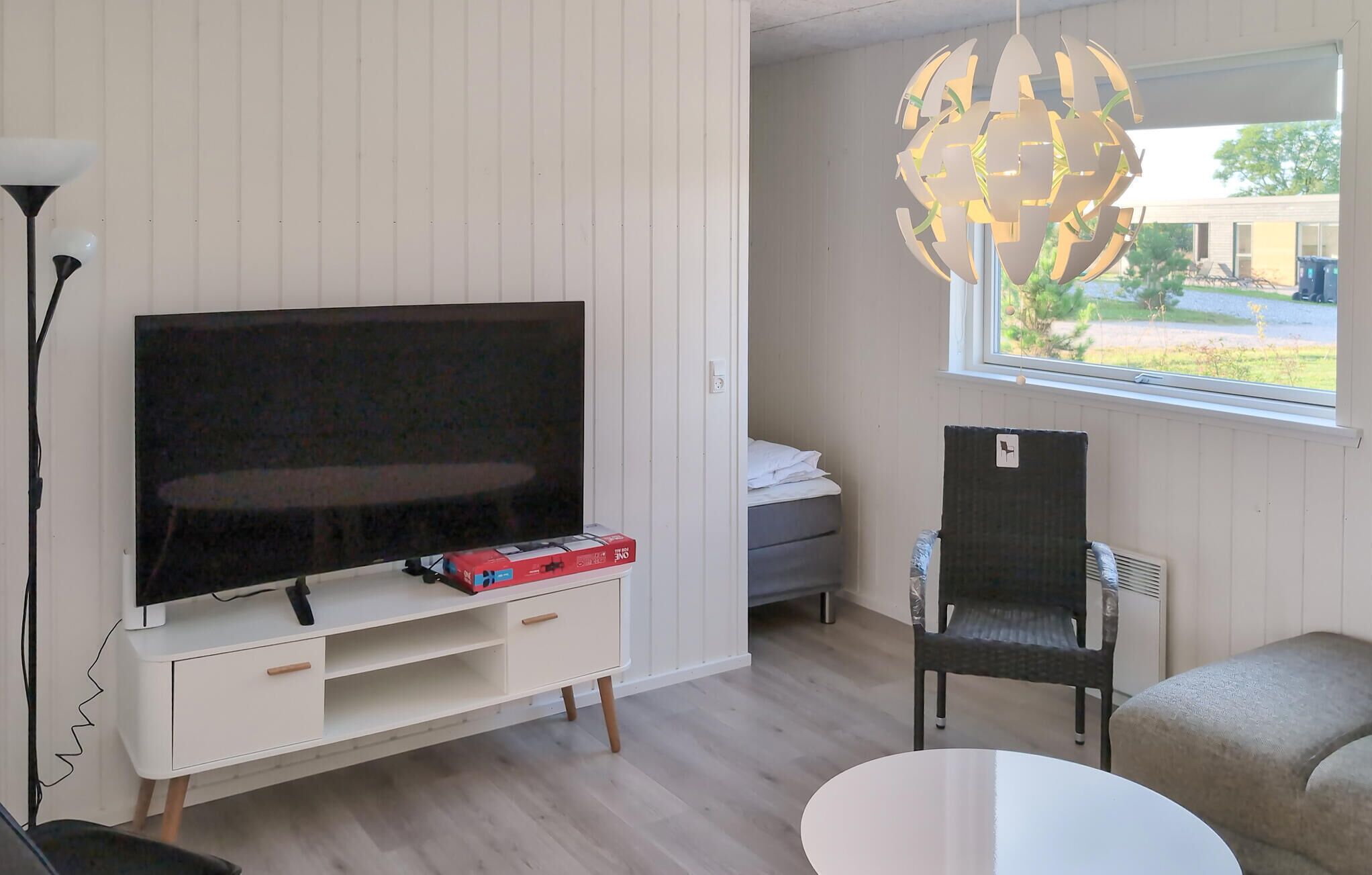 A modern living room with a flat-screen TV on a white TV stand, a dining area with a round table, a wicker chair, and a globe-shaped light fixture above a window.