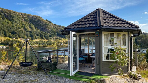A small gazebo with a glass door and windows, situated near a lake with a view of a forested mountain. - Stunning home in Korssund with WiFi (Korssund)