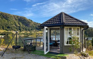 A small gazebo with a glass door and windows, situated near a lake with a view of a forested mountain. - Stunning home in Korssund with WiFi (Korssund)