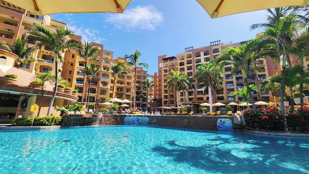 A hotel pool area with clear blue water, palm trees, and multiple balconies. -  2 Bedroom condo in Villa la Estancia in Nuevo Nayarit (Nuevo Nayarit)