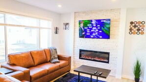 A modern living room with a leather sofa, a flat-screen TV mounted on a brick wall, a fireplace, and a small table. - New Modern & Luxury 3BR+2.5 Bath w/Garage Home in Saskatoon, SK (Saskatoon)