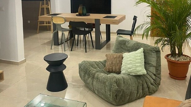 A modern living room with a dining area, a large black pendant light, a green sofa, a glass coffee table, and a potted plant.