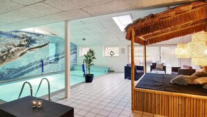 A hotel room with a large window, a bed, a small table with candles, a potted plant, and a view of a swimming pool. - 5 Star Holiday Home in Ebeltoft (Ebeltoft)