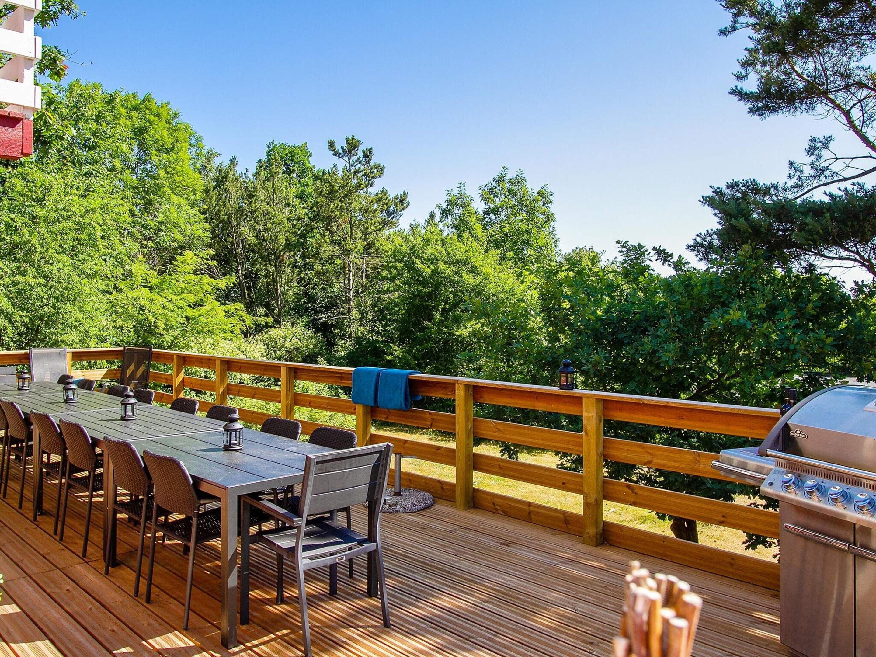 Terraza exterior con una mesa larga y sillas, rodeada de barandas de madera, con vista a un área boscosa.