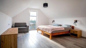 Ein Schlafzimmer mit schrägem Dach, einem Bett mit Holzrahmen, einem grauen Sofa, einem Holztisch und einem Fenster mit Jalousien.