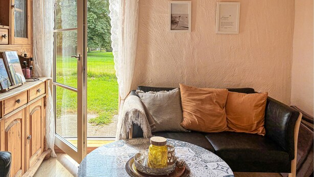 Une pièce chaleureuse avec un meuble en bois, un canapé en cuir noir agrémenté de coussins, une table ronde sur laquelle repose un plateau décoratif, et une vue sur un jardin verdoyant par une porte vitrée.