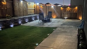 Un cortile con un patio in cemento, due sedie da giardino, un tavolino, una recinzione in legno e un lampione.