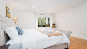 Un lit bien fait, avec du linge de lit blanc et un plaid beige, une lampe de chevet et un tableau encadré au mur.