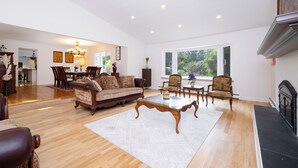Un salon spacieux avec un grand canapé, une table basse et plusieurs fauteuils. On aperçoit une cheminée et un coin repas au fond de la pièce.