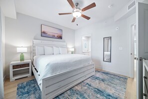 Ein Schlafzimmer mit einem Bett, einem Deckenventilator, einem Nachttisch, einer Lampe, einem Gemälde, einem Spiegel und einer Tür.