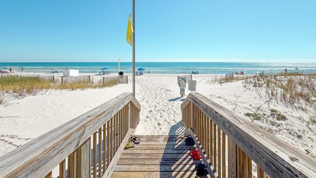 En træpromenade der fører til en sandstrand, med et flagstang og strandparasoller i baggrunden.