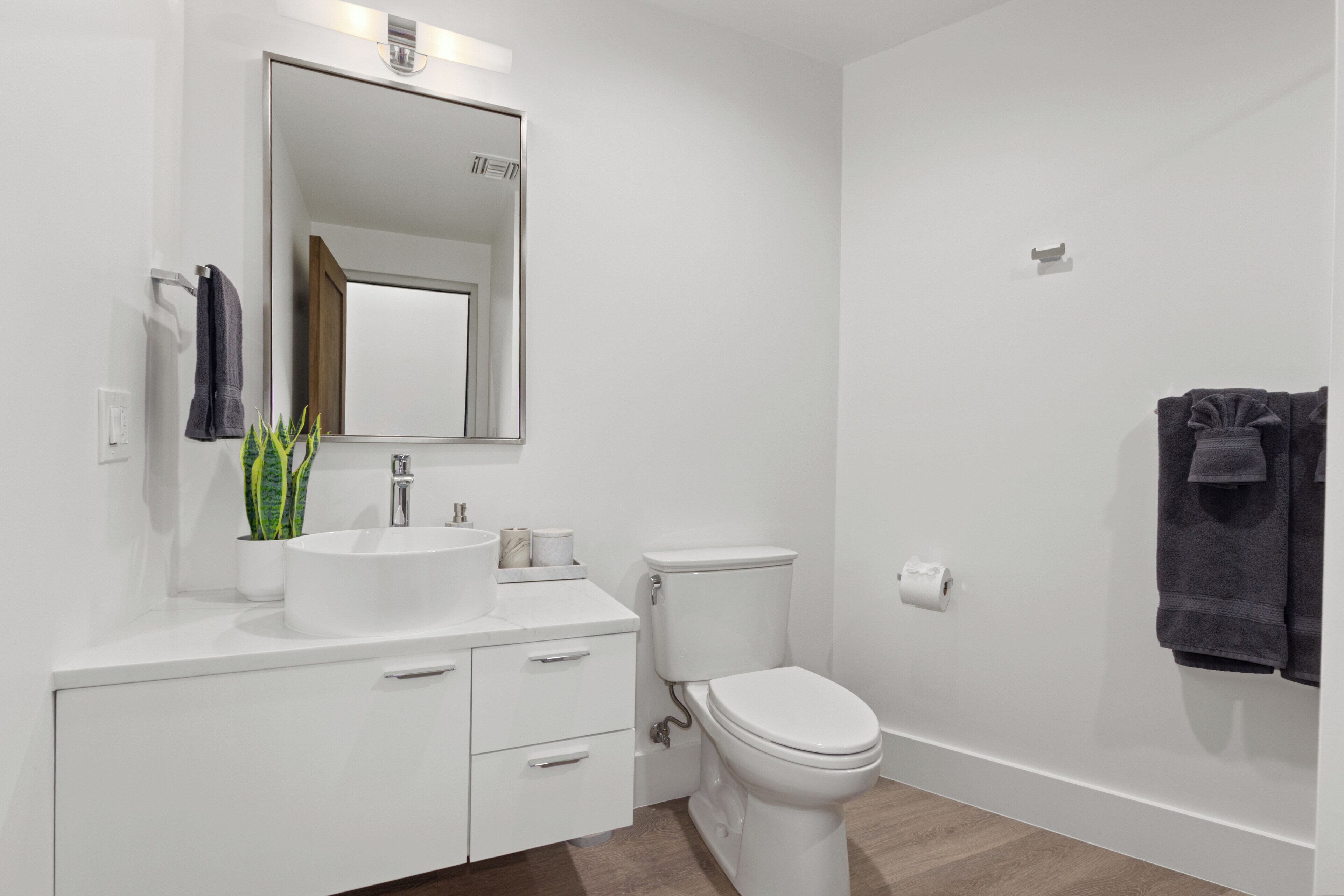 Un baño moderno con un lavabo blanco, espejo y retrete. Hay un perchero con toallas y una planta en maceta sobre el mostrador.