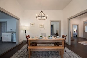 Un comedor con una mesa de madera dispuesta para cuatro personas, una araña y una cocina visible al fondo.