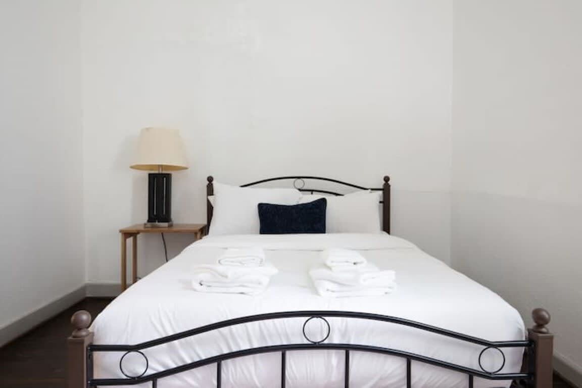 A neatly made bed with white linens and two folded towels, a bedside lamp, and a dark blue pillow.
