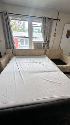 A room with a bed, a window with curtains, an air conditioning unit, and a small table. - 12th Ave The Bricks - Unit 5 (Birmingham)