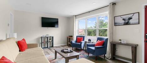 A modern living room with a beige sofa, blue armchairs, a coffee table, and a flat-screen TV mounted on the wall.