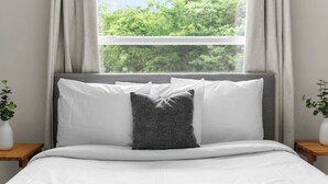 A neatly made bed with white and gray bedding, a view of trees through a window, and two bedside tables with plants. - Dugan Ave Dugout - Unit A (Birmingham)