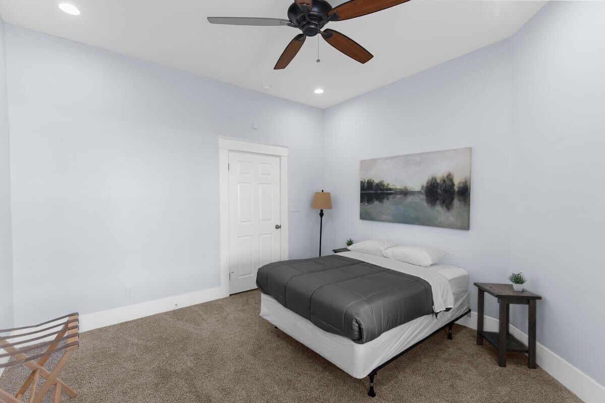 Un dormitorio con cama, ventilador de techo, puerta, lámpara, una mesita con una planta y un cuadro en la pared.