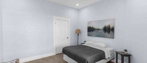 Un dormitorio con cama, ventilador de techo, puerta, lámpara, una mesita con una planta y un cuadro en la pared.