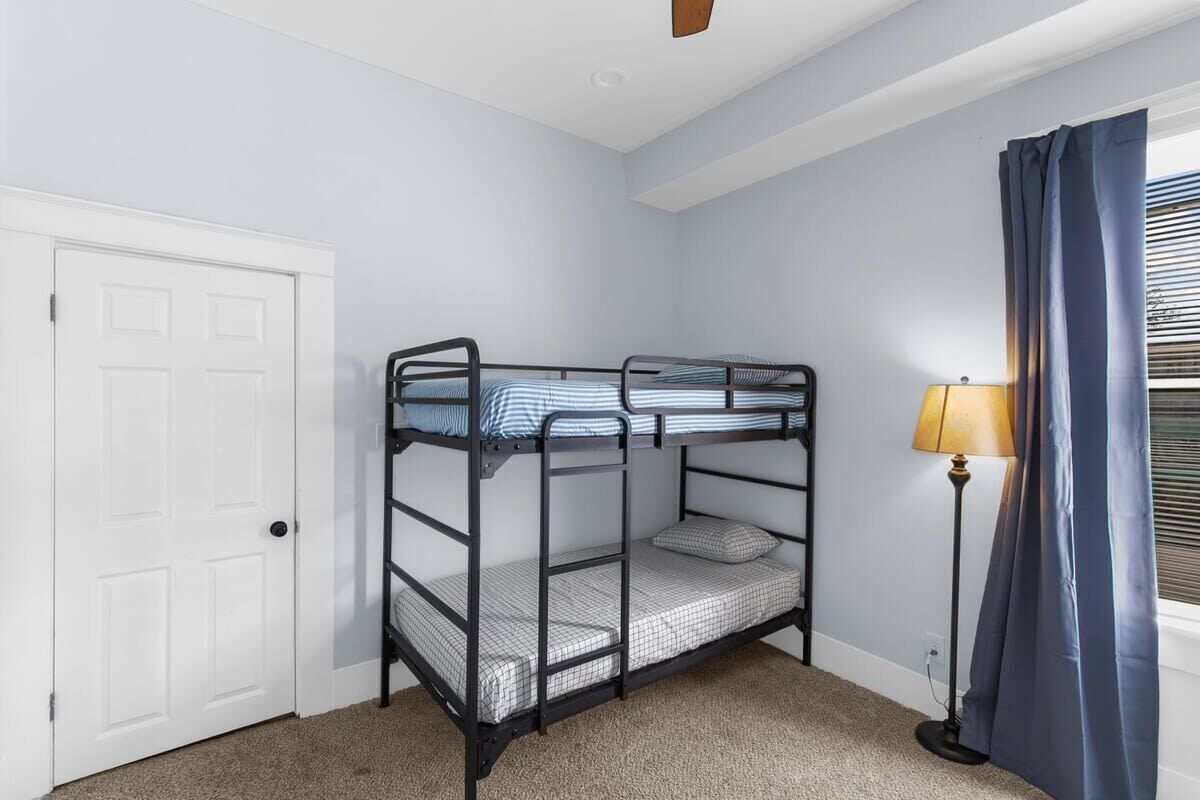 Quarto com beliche, porta branca, luminária de chão e janela com cortinas.
