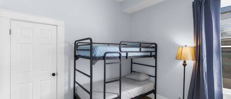 Quarto com beliche, porta branca, luminária de chão e janela com cortinas.