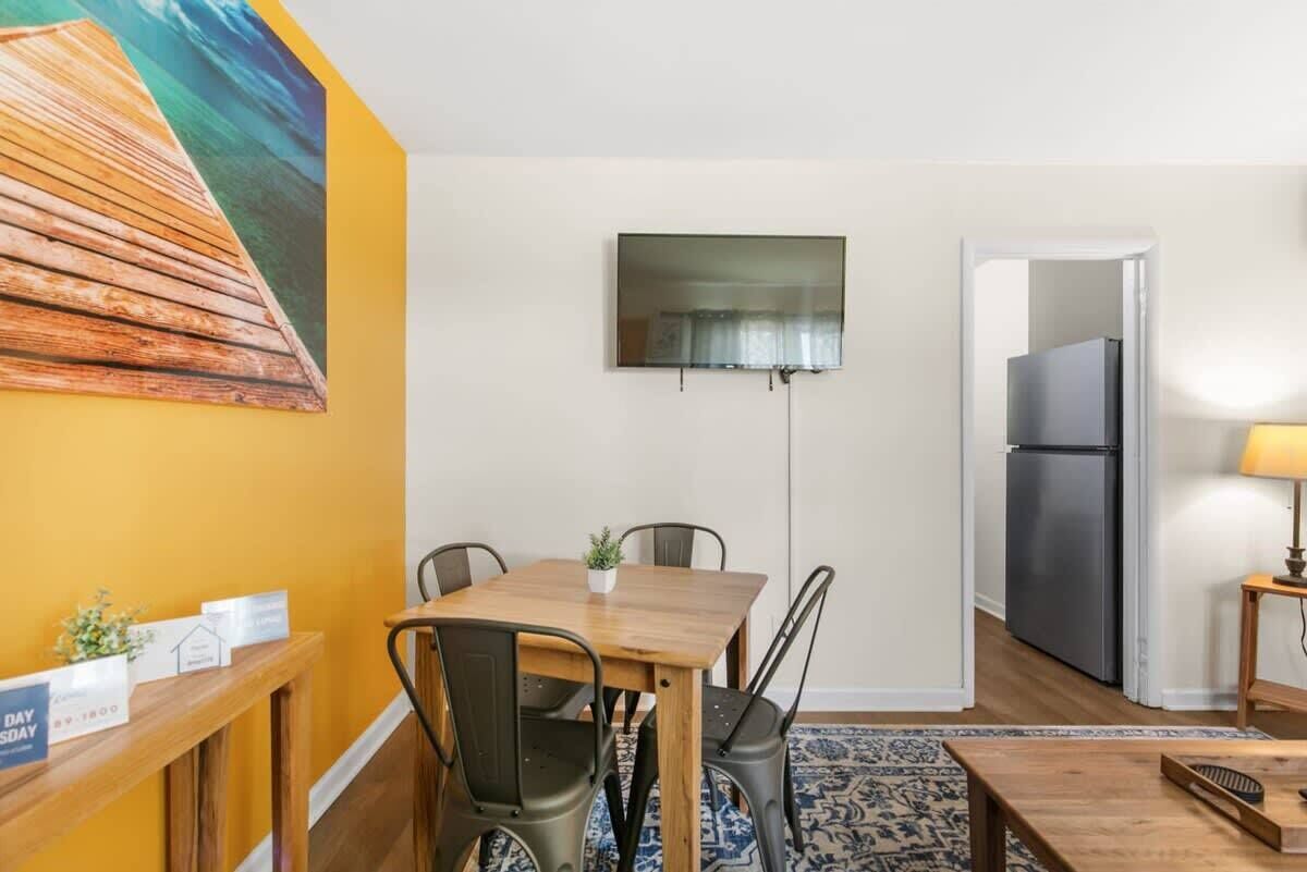 A compact living space with a dining area, a flat-screen TV mounted on the wall, a refrigerator, and a small table with a lamp.