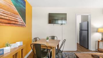 A compact living space with a dining area, a flat-screen TV mounted on the wall, a refrigerator, and a small table with a lamp.