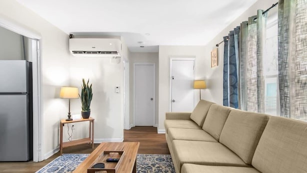 Ein modernes Wohnzimmer mit einem beigefarbenen Sofa, einem Holztisch und einem gemusterten Teppich. Ein kleiner Beistelltisch trägt eine Lampe und einen Topf mit einer Pflanze.