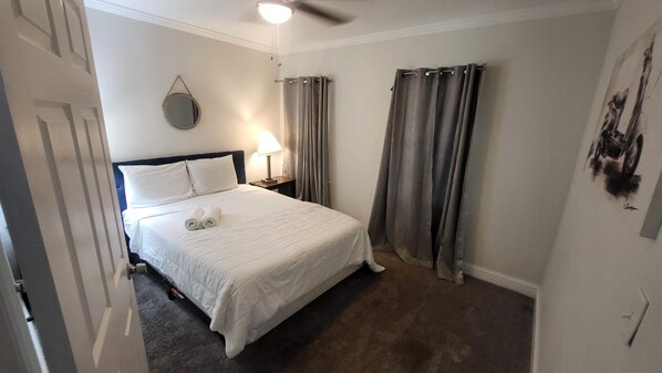 A neatly made bed with white linens and two rolled towels, a bedside lamp, and a ceiling fan in a room with a dark carpet and light-colored walls. - 83rd Street Comfy Cottage (Birmingham)