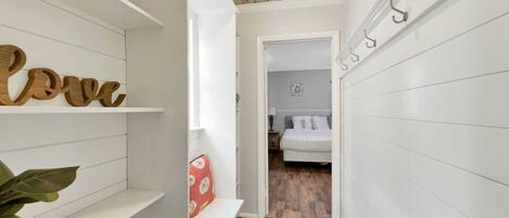 A narrow hallway with white walls and wooden flooring leading to a bedroom with a bed and a nightstand. Shelves on the left hold decorative items and a sign that reads "Love".