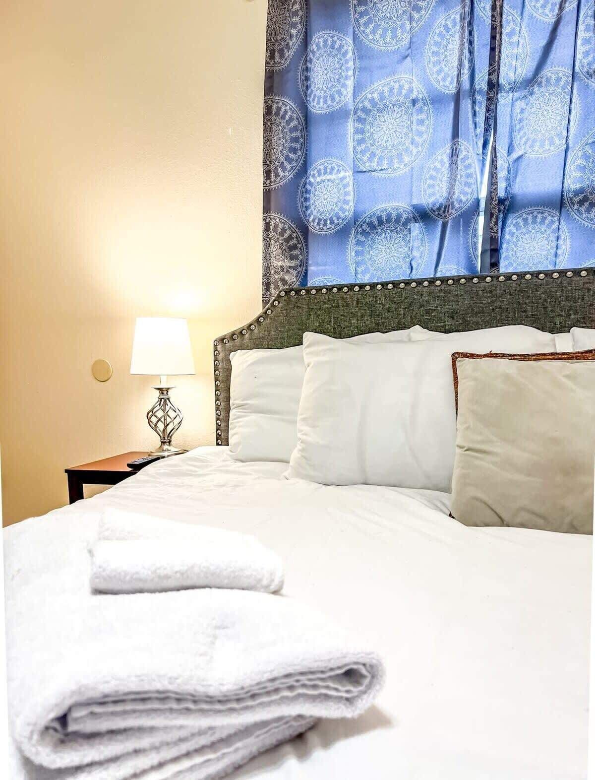 A neatly made bed with white linens and pillows, a bedside lamp, and a blue patterned curtain.