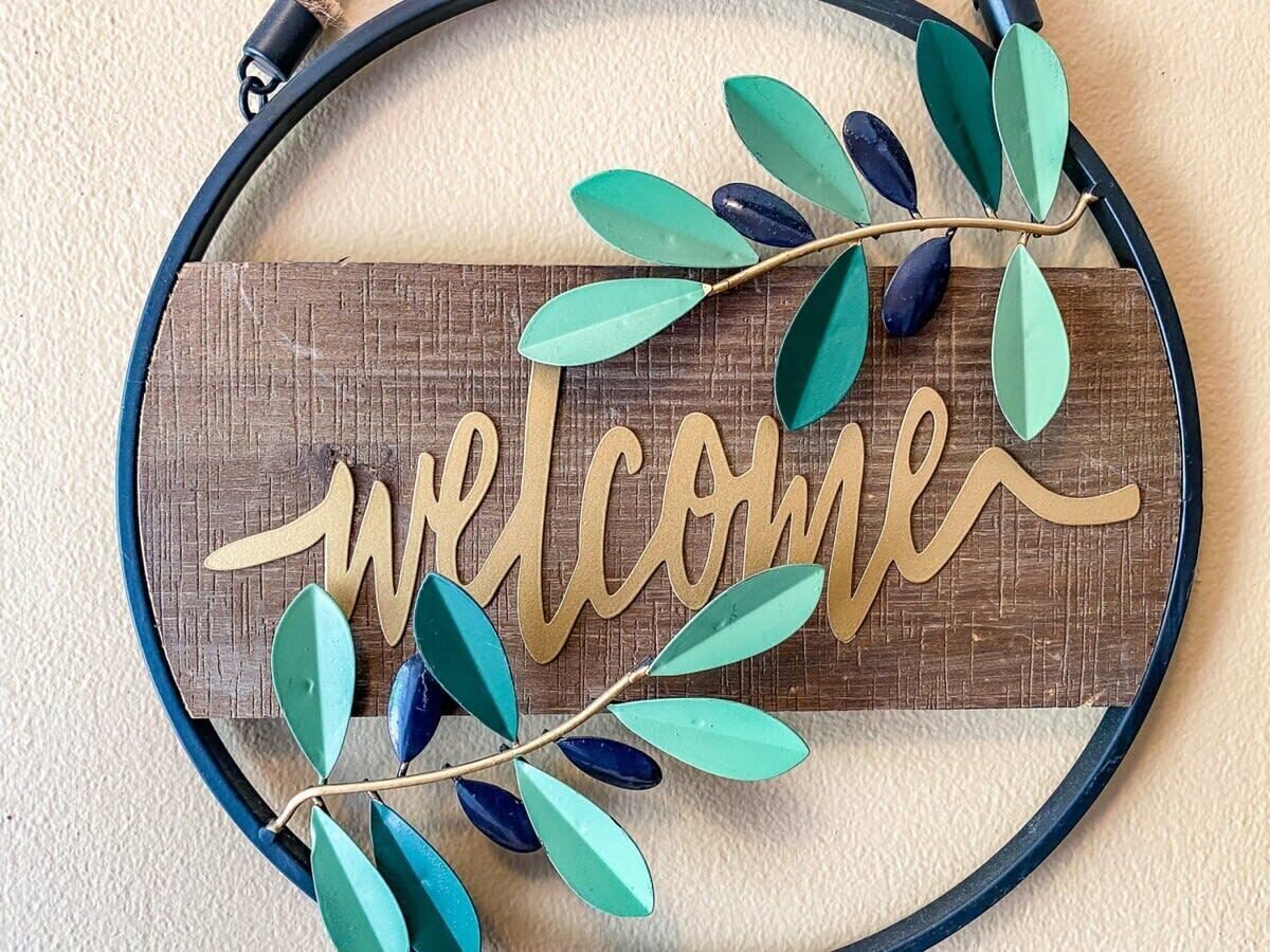 A wooden welcome sign with gold lettering, surrounded by green and blue leaves.