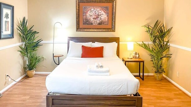 Una cama bien hecha con sábanas blancas y un cojín rojo decorativo, con dos plantas en macetas a cada lado, en una habitación con suelo de madera y una obra enmarcada sobre la cama.