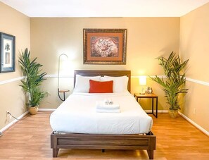 A neatly made bed with white linens and a red accent pillow, flanked by two potted plants, in a room with a wooden floor and a framed artwork above the bed. - Mahan Splashdown (Madison)