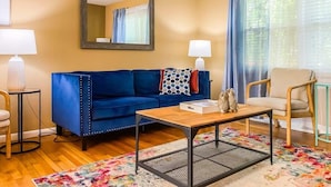 Una sala de estar con un sofá azul, una mesa de centro de madera, un espejo y una ventana con cortinas.